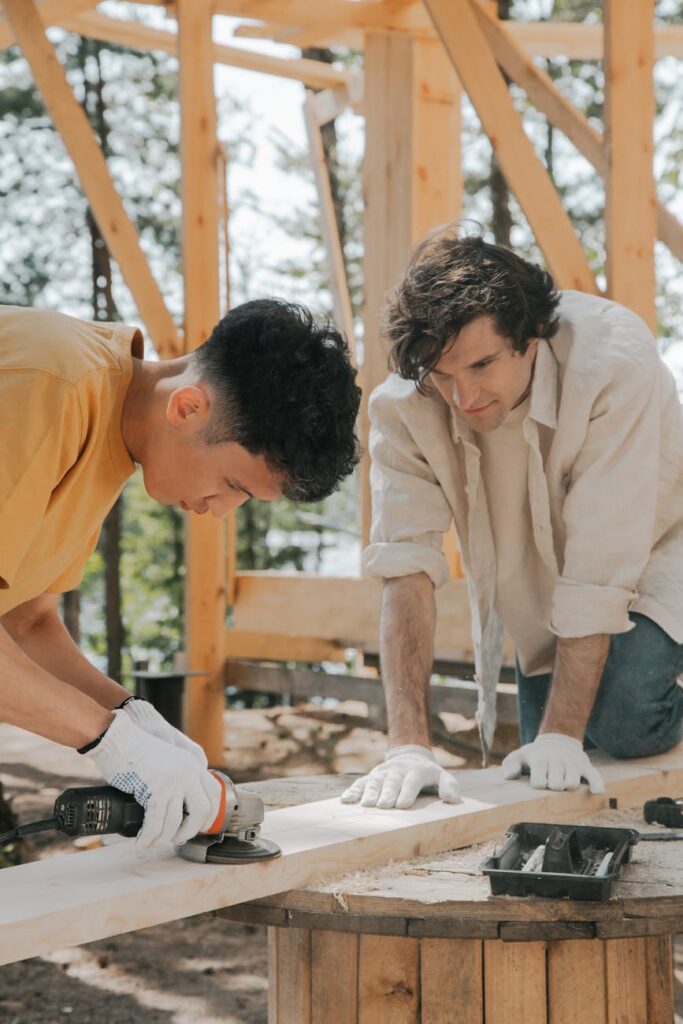 Two men collaborate to sand and polish a wooden beam at a construction site outdoors.