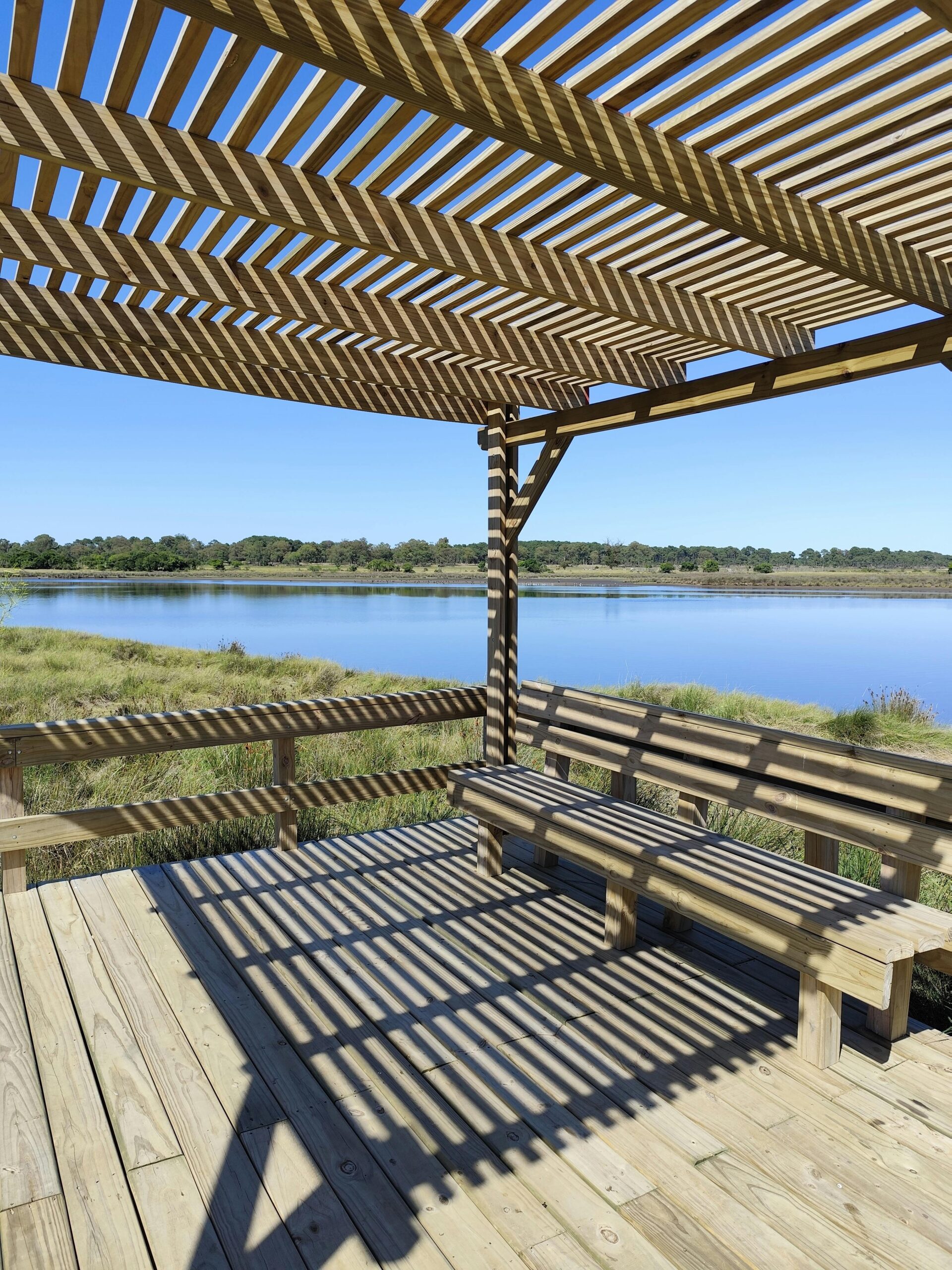 A wooden pergola casting shadows on a bench by a serene lake under a clear blue sky.